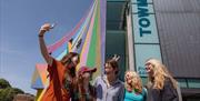 Five young people outside the Towner Gallery Eastbourne, the colourful exterior walls are behind them as they take a selfie.