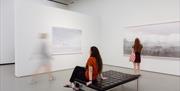 An internal photo of The Towner Gallery, Eastbourne, with people looking at artwork on the wall, one lady is sitting on a bench.
