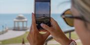Seaward view from outside the De La Warr Pavilion at Bexhill On Sea, a visitor's hands holding a mobile phone taking a photo of the gazebo, promenade,