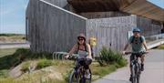 two cyclists passing the rye harbour discovery centre, a wooden buildng