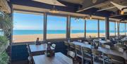 Interior of Perch on Lancing Beach a seaside restaurant with wooden tables and chairs, large windows overlooking a sandy beach and blue sea under a cl
