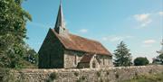 Small church with spire on the left side. It is framed by grass and trees with a stone wall in front.