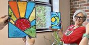 Two people in a cosy studio with brick walls and large windows holding up their finished pieces from their Stained Glass workshop: one features a colo
