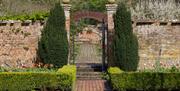 National Trust Bateman's topiary garden, wrought iron gate set within two pillars, a pair of trees and brick walls.  Path leading through the gates wi