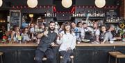 two people holding up their glasses at the bar with lots of people at the back..