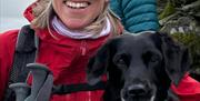 An image of a lady smiling in a red coat and winter hat holding walking sticks with a black dog next to her