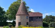 A traditional brick and timber Oast house with a conical roof, surrounded by bright yellow wildflowers and green trees under a clear blue sky.