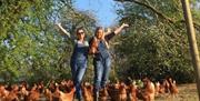 two women with arms high and smiling around lots of free roaming chickens
