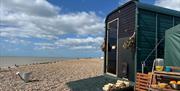 a horsebox sauna on Worthing's beach. The sky is blue with some clouds. There is a watering can on the pebbles, and to the right are shelves with wate
